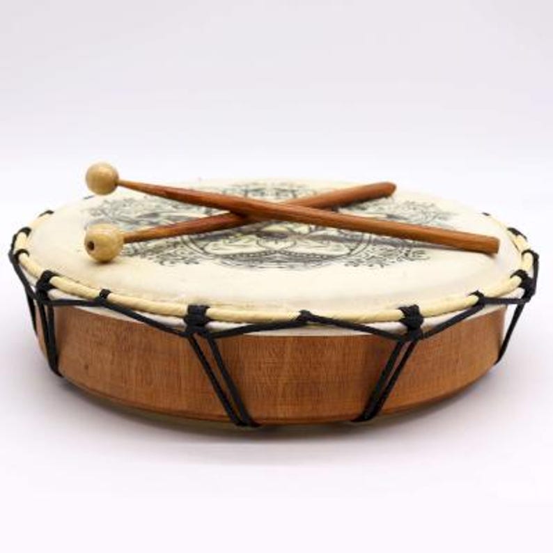 Op de afbeelding: Een kleine, ronde trommel met een lichtgekleurd vel met een zwart ontwerp. De trommel heeft een houten frame en is bespannen met zwart touw. Twee houten drumstokken met afgeronde uiteinden liggen erop.