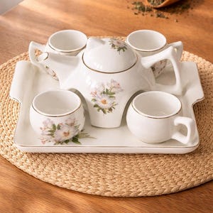 May include: A white porcelain tea set with floral accents, including a teapot, four teacups, and a serving tray. The set has gold trim and is arranged on a woven placemat. A wooden spoon with tea leaves is in the background.