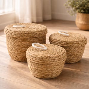 Peut inclure: Trois paniers de rangement ronds en matériau naturel tissé de couleur marron clair. Ils sont de tailles différentes et ont des couvercles avec des poignées en boucle blanches. Les paniers sont posés sur un sol en bois.