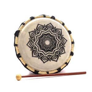 Op de afbeelding: Een kleine, ronde trommel met een cr&egrave;mekleurig vel met een zwart mandala-ontwerp. De trommel heeft een licht houten frame met zwarte accenten en een houten drumstok met een afgeronde kop.