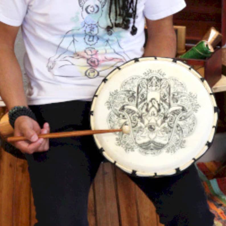 Op de afbeelding: Een ronde trommel met een cr&egrave;mekleurig oppervlak met een zwart Hamsa-handontwerp. De trommel heeft een zwarte rand en wordt bespeeld met een houten drumstok. De persoon draagt een wit t-shirt met een chakra-ontwerp.