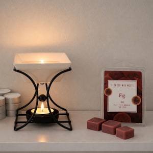 Può includere: Un bruciatore di cera nero e bianco con una candela accesa. Accanto, una confezione di cere profumate con l'etichetta "Fico" e tre cubetti di cera profumata. Diverse candele da tè impilate a sinistra.