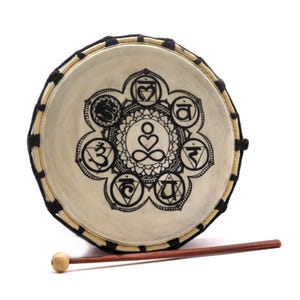 Op de afbeelding: Een ronde trommel met een natuurlijke huidkop, versierd met een zwart chakra-symboolontwerp. De trommel heeft een houten frame en wordt geleverd met een houten drumstok. Een muziekinstrument voor meditatie en spirituele praktijken.
