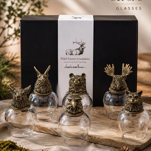 Peut inclure: Un ensemble de 6 verres avec des bases en métal couleur bronze antique en forme de têtes d'animaux, incluant un sanglier, un lapin, un ours, un aigle, un élan et un renard. Les verres sont présentés devant une boîte noire marquée Wild Forest Creatures.