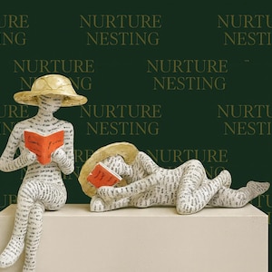 Peut inclure: Deux sculptures blanches de personnages lisant des livres, l'un assis et l'autre allongé. Les deux personnages portent des chapeaux et sont recouverts de texte. Les personnages sont sur une plateforme blanche sur fond vert foncé avec "Nurture Nesting".