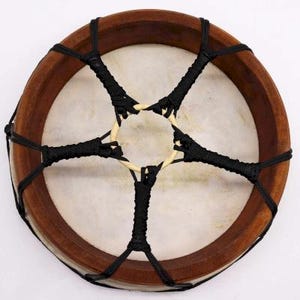 Op de afbeelding: Een ronde, handgemaakte trommel met een lichtgekleurd, gespannen huidoppervlak. Het houten frame van de trommel is donkerbruin en heeft een stervormig patroon van zwart touw over het oppervlak. Muziekinstrument.
