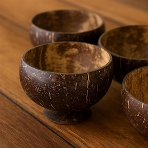 May include: Four dark brown coconut bowls with a natural, textured finish. One bowl is centered, showing its pedestal base. The bowls are arranged on a wooden surface. Each bowl is made from a coconut shell.