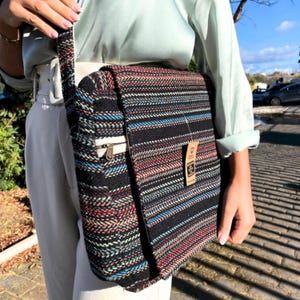 Op de afbeelding: Een zwarte en veelkleurige gestreepte messenger bag met een klepsluiting. De tas heeft een witte ritszak en een bruin label. De tas is gemaakt van geweven stof en heeft een lange riem.