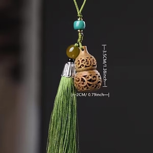 Pode incluir: Um pingente de madeira em forma de cabaça com desenhos entalhados intrincados, medindo 2 cm de largura e 3,5 cm de altura. Possui uma borla verde, uma conta turquesa e um cordão verde. O pingente é um acessório decorativo.