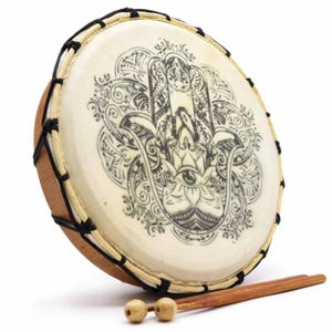 Op de afbeelding: Een ronde trommel met een cr&egrave;mekleurig vel met een zwart Hamsa-handontwerp. De trommel heeft een houten frame en is geregen met zwart touw. Twee houten drumstokken met afgeronde koppen liggen voor de trommel.