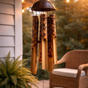 May include: A bamboo wind chime with a dark brown coconut shell top and cylindrical chimes. The chimes have a natural wood colour with dark brown patterned designs. The wind chime hangs from a black string, with a small wooden piece at the bottom.