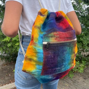 May include: A tie-dye drawstring backpack in a vibrant mix of colours, including yellow, orange, red, blue, and purple. It has a front zippered pocket and cream-coloured rope drawstrings.