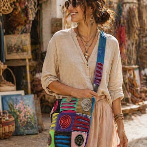 May include: A patchwork shoulder bag with a blue strap. The bag is made up of squares in various colours, including pink, purple, green, and blue, with embroidered spiral and circular designs. The woman is wearing a beige shirt and pink skirt.