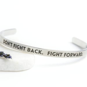 May include: Silver bangle bracelet with the inscription "Don't fight back. Fight forward."