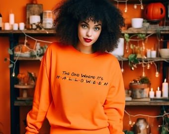 Op de afbeelding: Fel oranje sweatshirt met de zwarte tekst "The One Where It's Halloween". Een persoon met donker krullend haar en rode lippenstift draagt het sweatshirt. De achtergrond bevat Halloween-decoraties.
