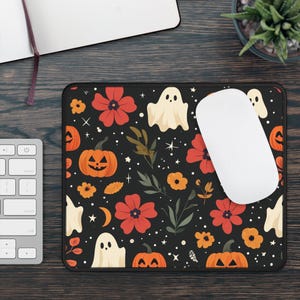 Może przedstawiać: Czarna podkładka pod mysz z motywem Halloween. Wzór przedstawia pomarańczowe dynie, czerwone kwiaty, białe duszki i zielone liście na czarnym tle. Biała mysz komputerowa spoczywa na podkładce.