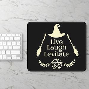 May include: Black rectangular mousepad with the words "Live Laugh Levitate" in a cream-colored font. The design includes a witch hat, two broomsticks, a pentagram, and a laurel wreath. A white keyboard is visible on the left.