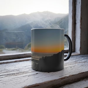 May include: A black coffee mug with a sunset over the ocean scene printed on it. Steam is rising from the mug, which is sitting on a wooden windowsill.