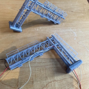 May include: Two gray 3D printed models of a bridge structure with wires attached. The models are on a wooden surface.
