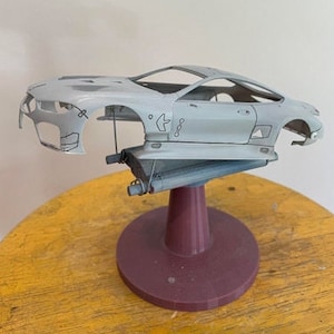Può includere: Una carrozzeria di auto modello grigio chiaro montata su un supporto bordeaux. Il design dell'auto è visibile, con aree aperte per finestre e dettagli interni. Il supporto è su una superficie gialla.