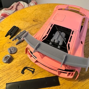 Puede incluir: Un coche modelo rosa con un alerón trasero gris, un motor negro y varias piezas dispuestas sobre una superficie de madera.