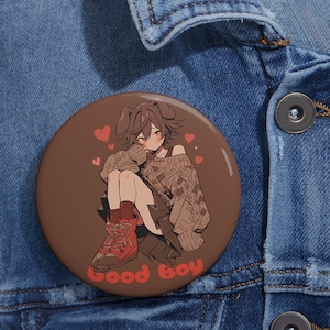 May include: A brown button with a cartoon illustration of a person with brown hair wearing a brown sweater and red boots. The person is sitting with their legs crossed and has a heart-shaped nose. The text "Good Boy" is written in a retro font at the bottom of the button.