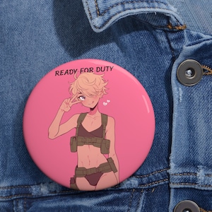May include: A pink button with the text "READY FOR DUTY" in black. The button features a cartoon illustration of a person with blonde hair wearing a green and brown military-style outfit. The person is smiling and giving a peace sign.