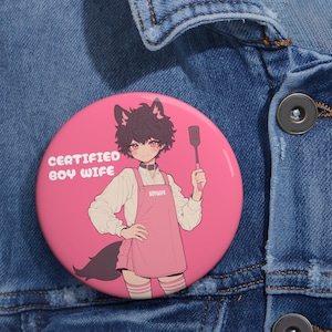 May include: A pink button with a cartoon illustration of a person with black hair and cat ears wearing a pink apron and holding a spatula. The text "CERTIFIED BOY WIFE" is printed on the button.