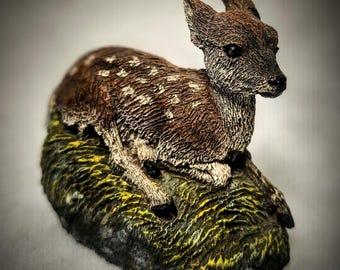 May include: A small, brown and white ceramic figurine of a fawn lying down on a green and brown base.