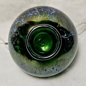 May include: A round, iridescent glass object with a dark green interior and a silver rim. The exterior has a mottled appearance with shades of green, gold, and blue. The object appears to be a decorative piece or a container.