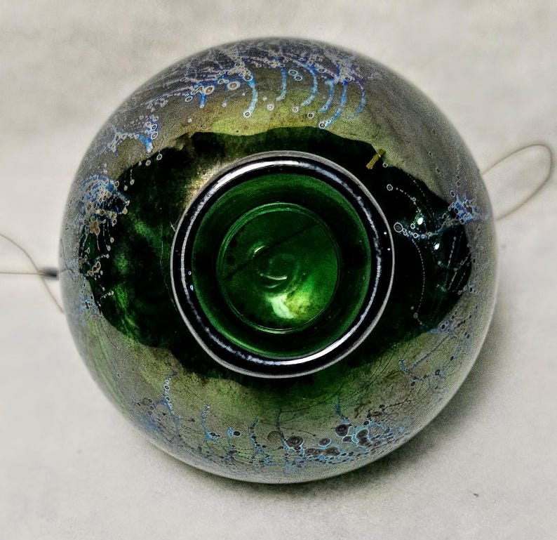 May include: A round, iridescent glass object with a dark green interior and a silver rim. The exterior has a mottled appearance with shades of green, gold, and blue. The object appears to be a decorative piece or a container.