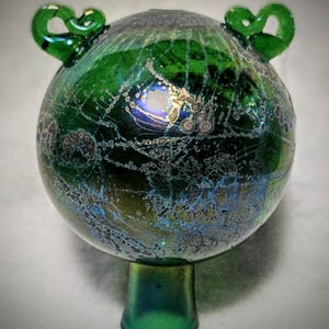 May include: A handcrafted, iridescent green glass orb with a flared base and two decorative loop handles. The glass has a textured, speckled appearance with hints of blue and gold. The decorative piece is likely a vase or art object.