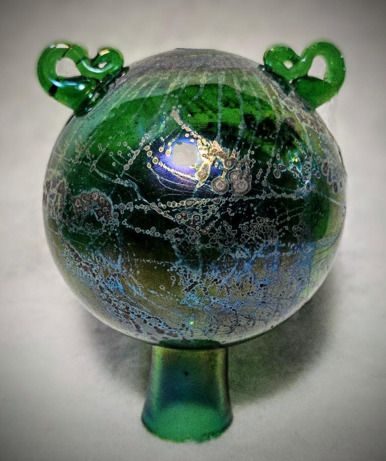 May include: A handcrafted, iridescent green glass orb with a flared base and two decorative loop handles. The glass has a textured, speckled appearance with hints of blue and gold. The decorative piece is likely a vase or art object.