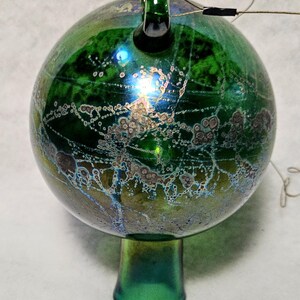 May include: A green glass ornament with a round shape and a small stem. The ornament has a textured surface with iridescent colors and a loop at the top. A wire extends from the top.