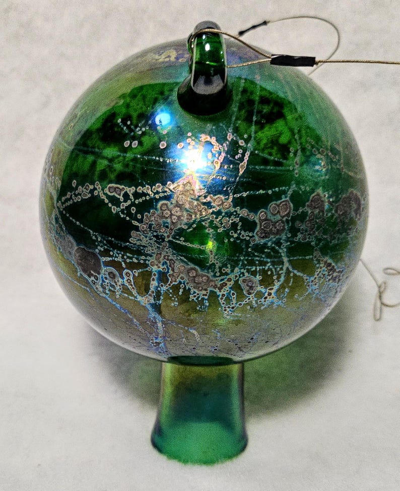 May include: A green glass ornament with a round shape and a small stem. The ornament has a textured surface with iridescent colors and a loop at the top. A wire extends from the top.