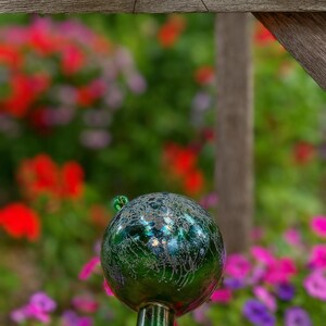 May include: A decorative green glass gazing ball with a crackle effect, mounted on a matching stem. The orb is set against a blurred background of colorful flowers, including red, pink, and purple blooms.