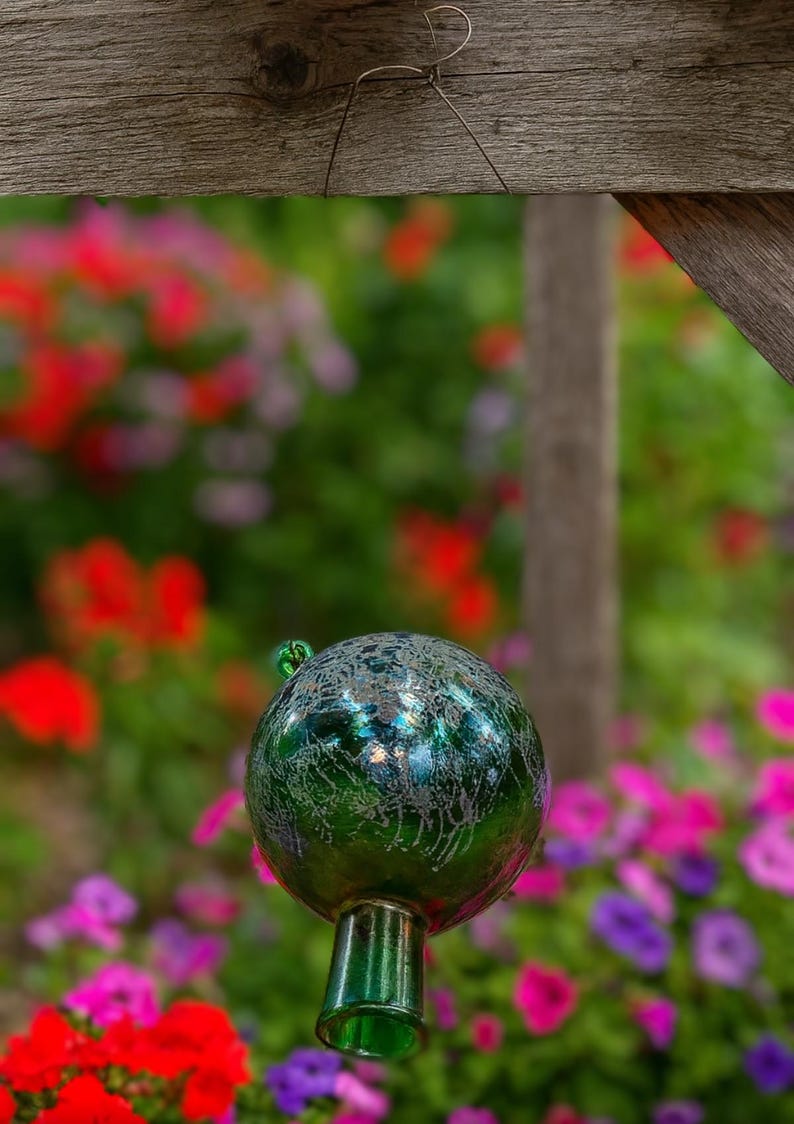May include: A decorative green glass gazing ball with a crackle effect, mounted on a matching stem. The orb is set against a blurred background of colorful flowers, including red, pink, and purple blooms.