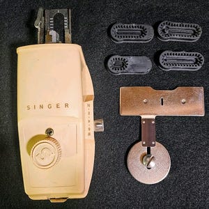Può includere: Un set di accessori per macchina da cucire Singer vintage. Il set include una testa della macchina color crema con il logo Singer, diversi piedini neri e una piastra metallica con una base circolare. Gli oggetti sono disposti su una superficie scura.