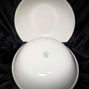 Puede incluir: Dos cuencos blancos Corelle Vitrelle, uno invertido para mostrar la parte inferior con el nombre de la marca y la información del producto. Los cuencos son redondos con un acabado liso y brillante. El fondo es una tela oscura y texturizada.
