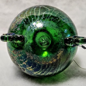 May include: A round, green glass orb with a textured surface and two small, dark green glass loops attached. The orb has a swirling pattern of green, blue, and gold colors. A thin wire extends from each loop.