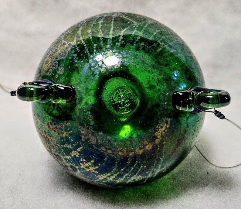 May include: A round, green glass orb with a textured surface and two small, dark green glass loops attached. The orb has a swirling pattern of green, blue, and gold colors. A thin wire extends from each loop.