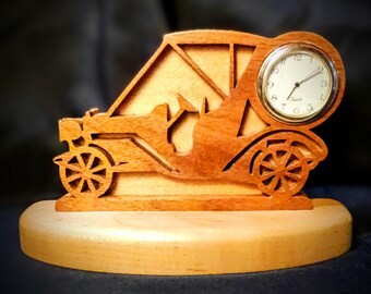 May include: A wooden clock in the shape of a vintage car. The clock has a round face with black numerals and hands. The car is made of light brown wood and sits on a light-colored wooden base. The wheels are detailed with spokes.