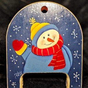 Puede incluir: Decoración de madera de un muñeco de nieve sobre un fondo azul con copos de nieve blancos. El muñeco de nieve lleva un gorro amarillo, una bufanda roja y mitones rojos. El nombre "Patsy" está escrito en cursiva.