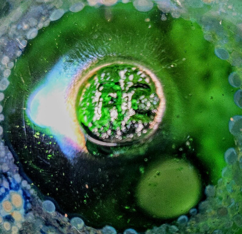 May include: Abstract macro shot of a glass object with a central green core surrounded by a dark green and blue-tinged border. The core has a textured, bubbly appearance, with iridescent light refracting through the glass.