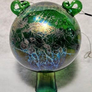 May include: A decorative glass ornament with a round body and a green stem. The ornament is primarily green with iridescent blue and gold accents. Two green loops are attached to the top. A wire extends from the ornament.