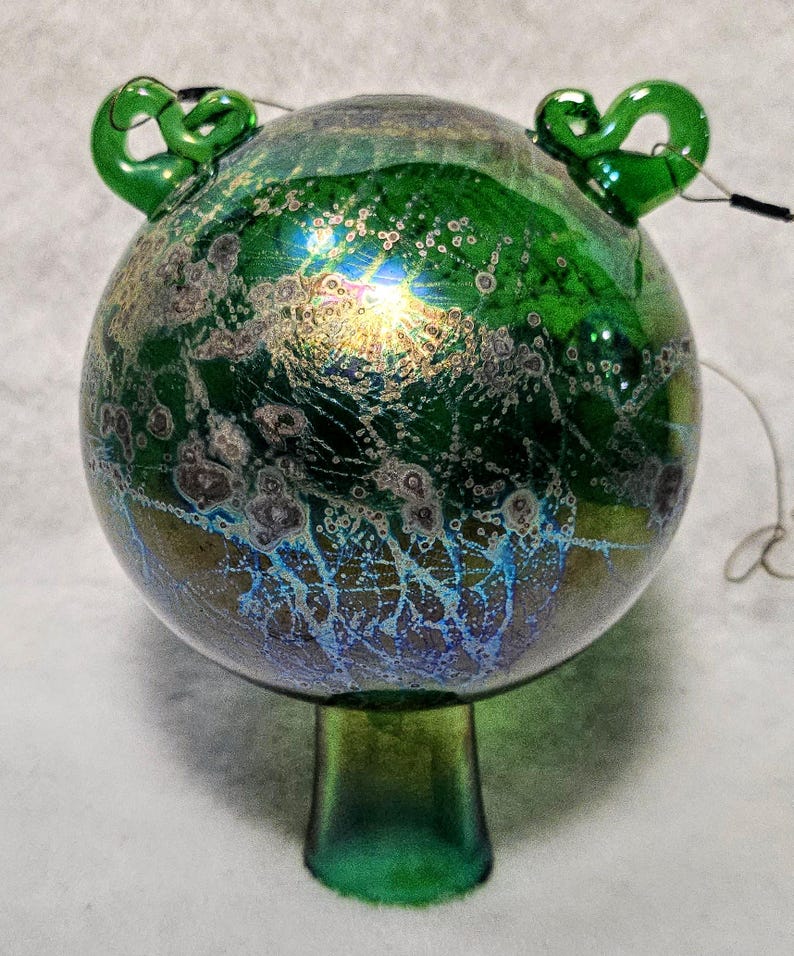 May include: A decorative glass ornament with a round body and a green stem. The ornament is primarily green with iridescent blue and gold accents. Two green loops are attached to the top. A wire extends from the ornament.