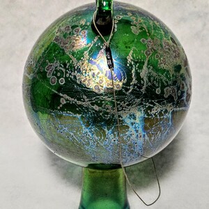 May include: A green glass ornament with a round shape and a small green stem. The ornament has a textured surface with a mix of green, blue, and gold colors. A wire is attached to the top of the ornament.