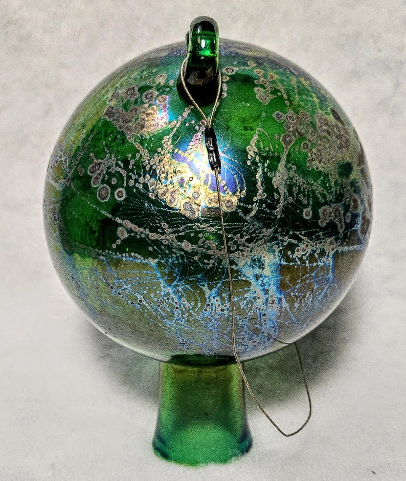 May include: A green glass ornament with a round shape and a small green stem. The ornament has a textured surface with a mix of green, blue, and gold colors. A wire is attached to the top of the ornament.