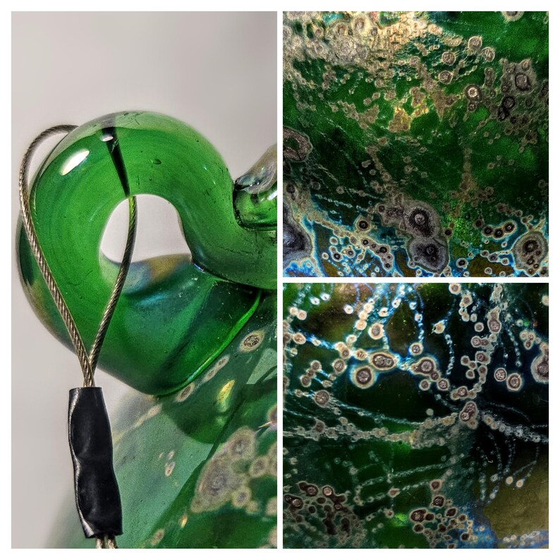 May include: Close-up of a green glass sculpture with a loop and a wire attached. The glass has a textured surface with white and brown spots. The sculpture is on a white background. The sculpture is a decorative art piece.