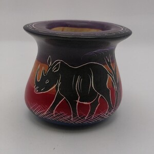 May include: A small, hand-painted ceramic pot with a purple, red, orange, and blue design. The pot features a black silhouette of a rhinoceros walking through a savanna landscape.